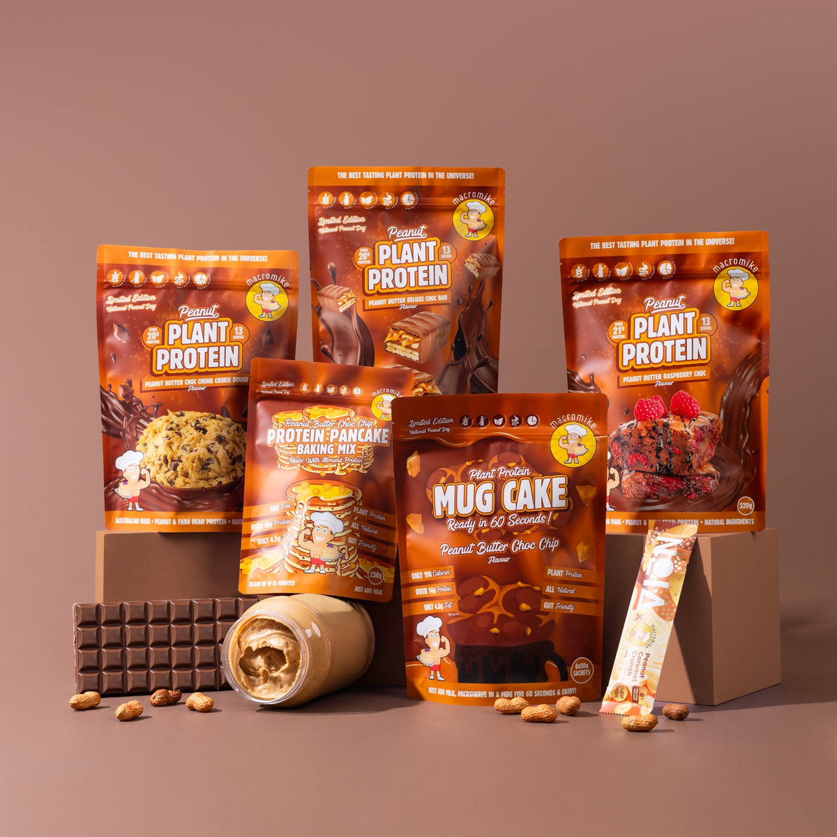 A group of Plant Based Protein snack products, including various pouches of protein powder, a protein pancake, a protein mug cake, and a protein bar, arranged on display blocks with chocolate and peanut props against a solid brown background.